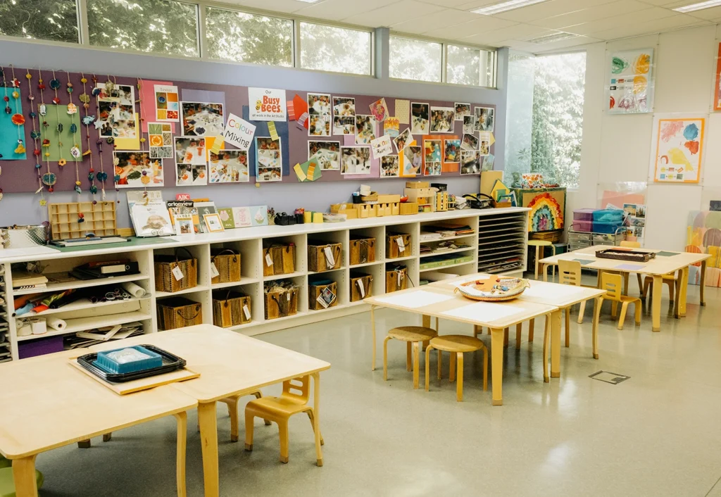 A well-organized childcare classroom with low tables, chairs, shelves filled with art supplies, and colorful artwork displayed on the wall. Bright windows let in natural light.