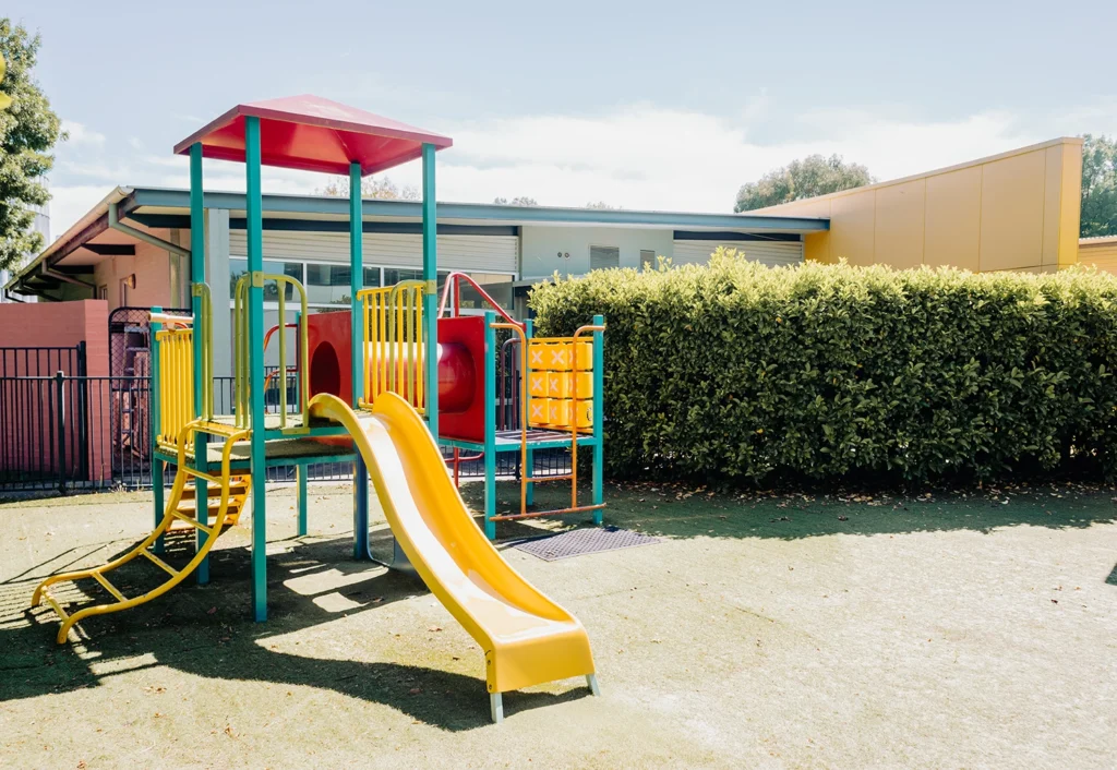 A brightly colored playground structure with a yellow slide, climbing bars, and a red roof stands on a grassy area near the day care building, partially enclosed by a hedge.