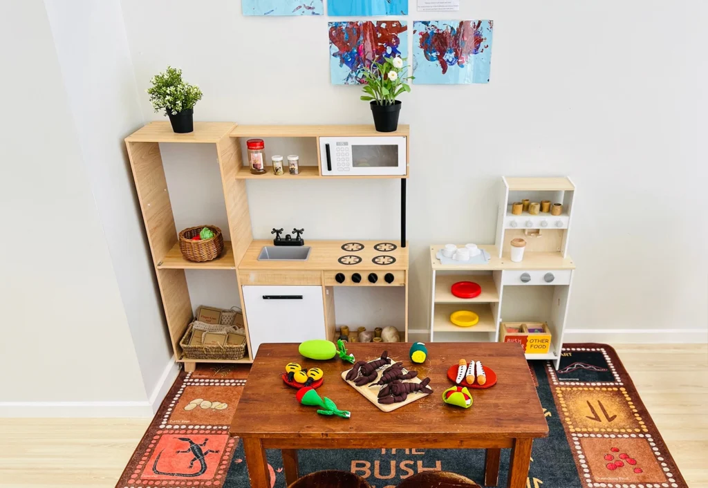 A small children's play kitchen perfect for a kindergarten or childcare setting, featuring wooden shelves, a toy microwave, a toy stove, various play food items, and a wooden table with chairs on a colorful rug.