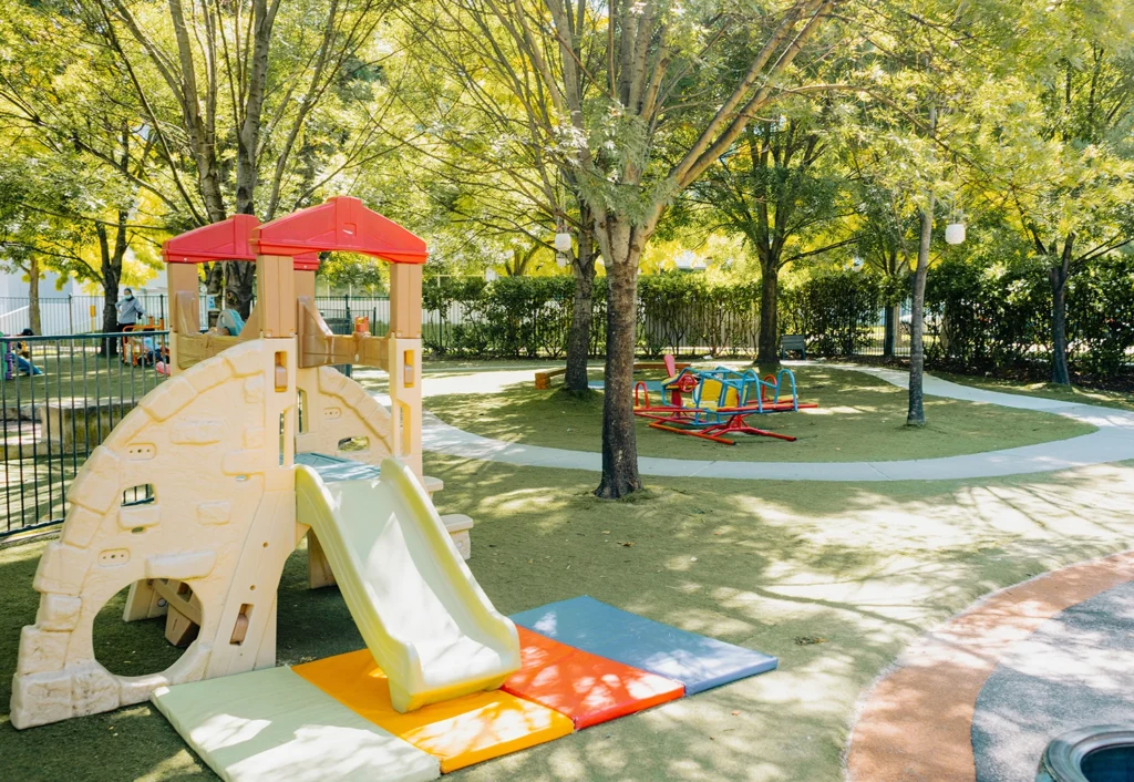 A playground perfect for a preschool features a plastic slide, a cushioned mat at its base, and a red merry-go-round in a shaded area with trees and a surrounding fence.