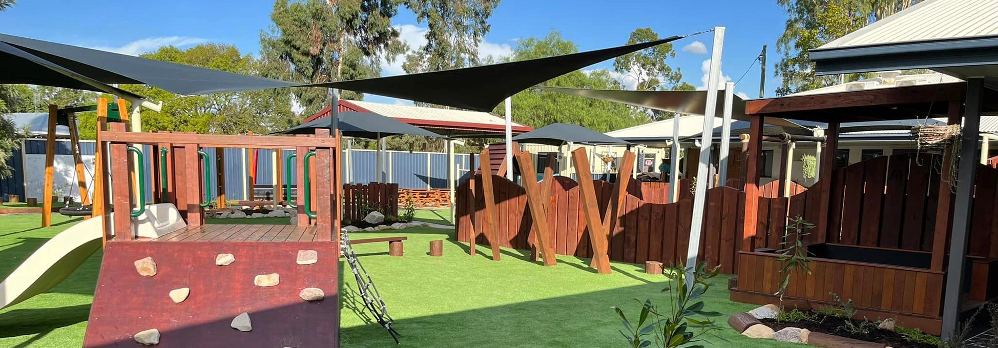 A well-equipped outdoor play area at Busy Bees Chinchilla daycare features shade sails, climbing structures, a slide, surrounded by wooden fencing and nearby trees.