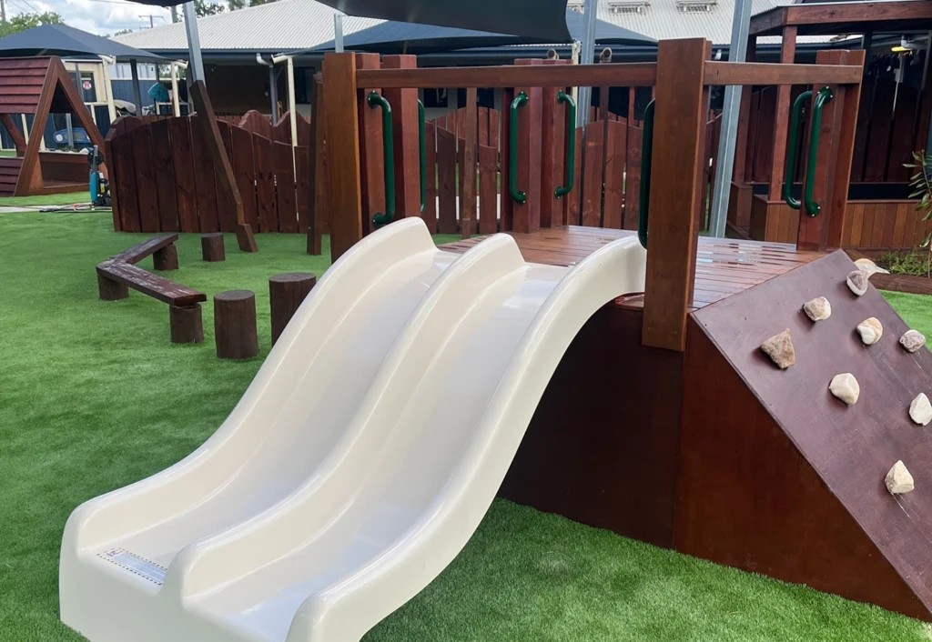 A playground at Chinchilla Kindergarten featuring a white double slide, a wooden climbing structure, and log stepping stones for balance play.