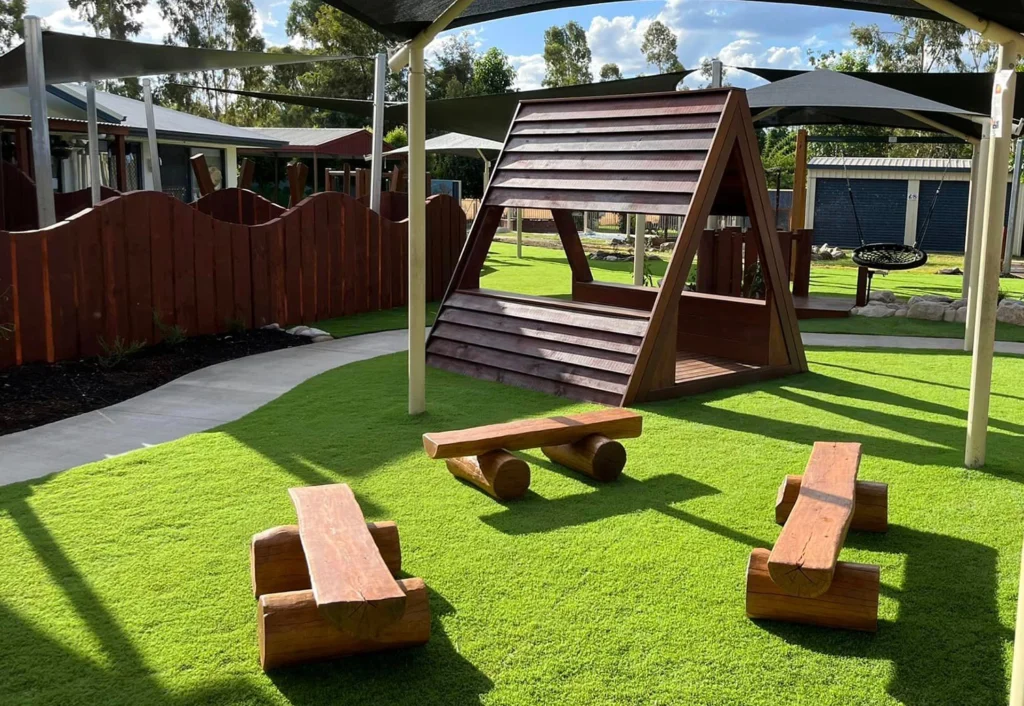 Outdoor play area at Chinchilla Kindy with artificial grass, wooden triangular playhouse, and wood benches. Modern shade structures and wooden fence surround the area. Trees and sky visible in the background.