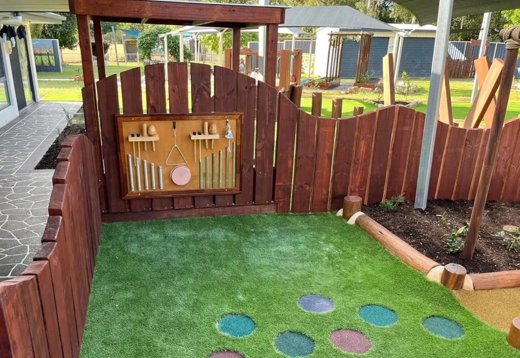 A playground corner at Chinchilla childcare with a wooden fence, musical panel wall, artificial turf, and colorful stepping stones. Nearby is a garden bed.