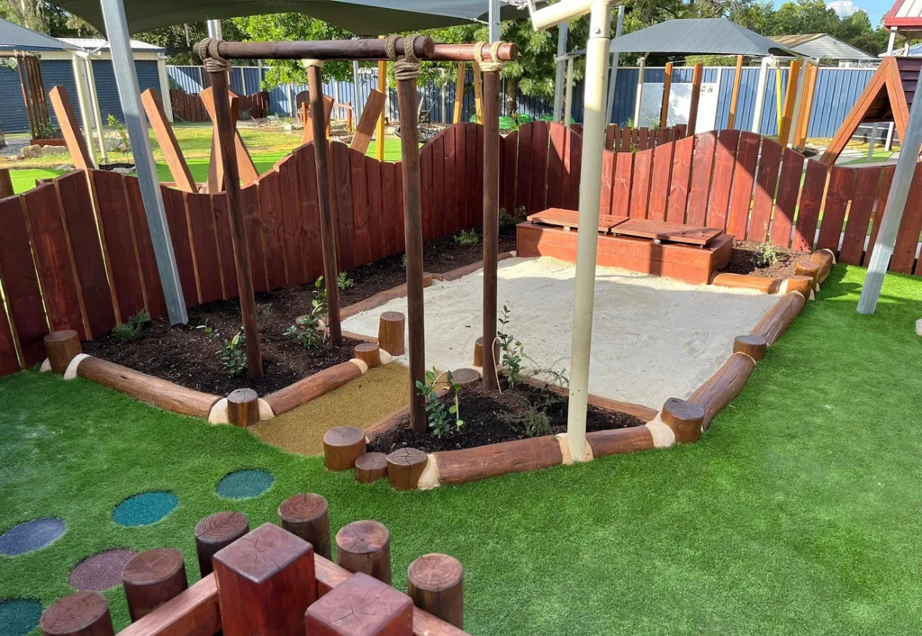 A sandpit in the nursery playground at Chinchilla day care centre surrounded by green artificial turf, a wooden fence and various timber play structures in the background.