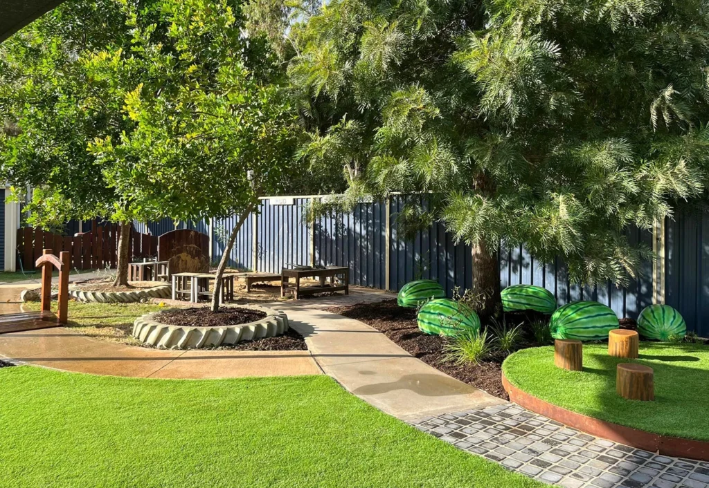 Outdoor playground at Busy Bees Chinchilla daycare with green grass, trees, a bike bath, and decorative objects painted to look like watermelons.