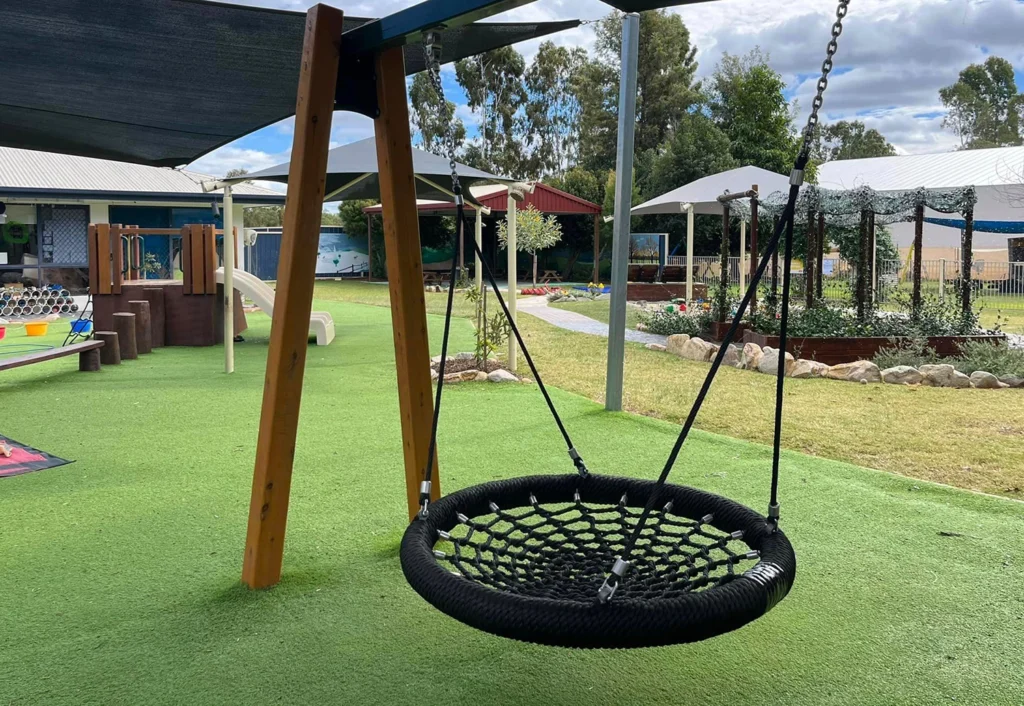 A children's playground at Chinchilla day care centre featuring a rope nest swing on a wooden frame. In the background are various play structures and a garden.