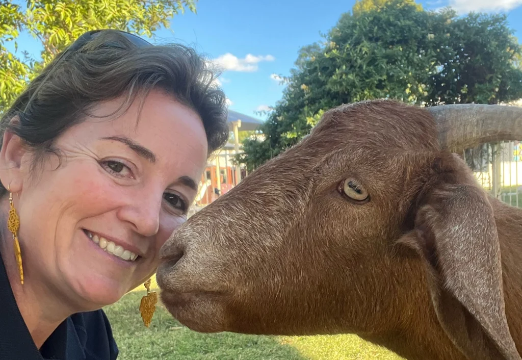 An Early Childhood Educator is smiling at the camera, while a goat stands very close to their face. Trees and the Chinchilla childcare building are visible in the background.