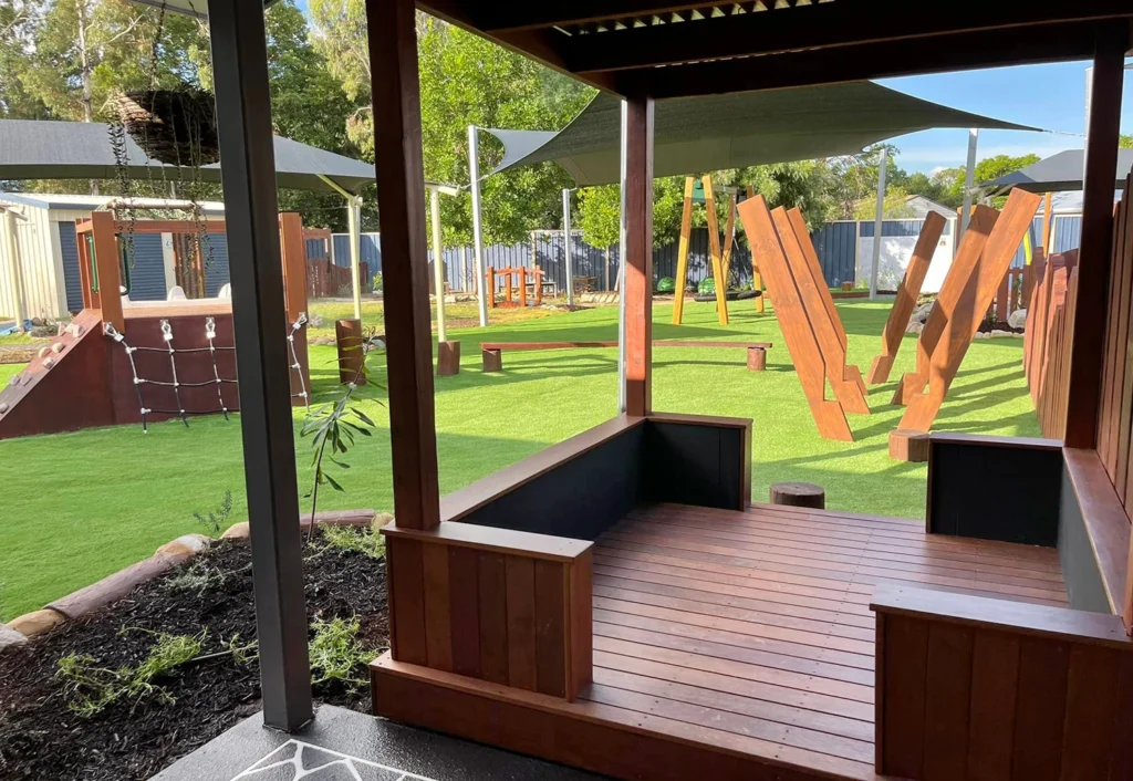 A playground with wooden climbing structures, balance beams, and shaded areas is seen from a wooden gazebo at Chinchilla Kindy.