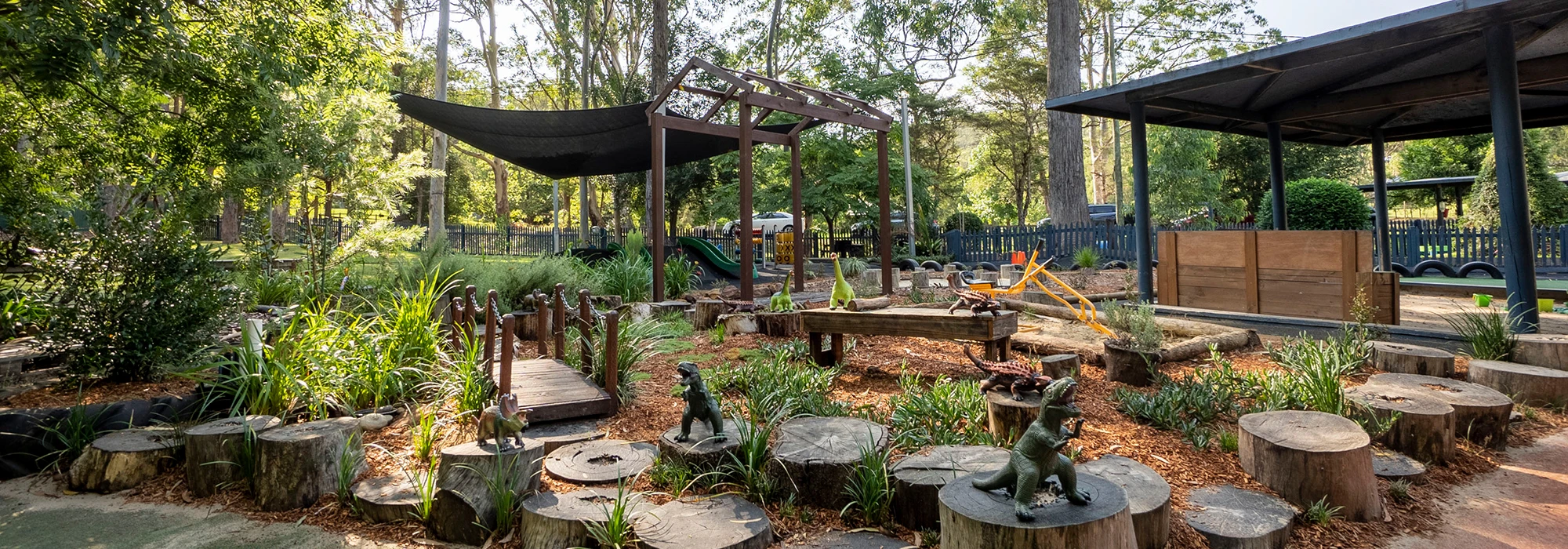 Outdoor playground at Fountaindale preschool featuring a timber bridge and log stepping stones, surrounded by trees and plants. The background includes shaded areas and a sandpit with a toy excavator, creating an adventure for the little ones.