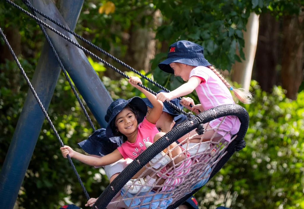 Children smiling as they play on a circular rope swing at Follyfoot Farm preschool located on the Central Coast. Trees are visible in the background.