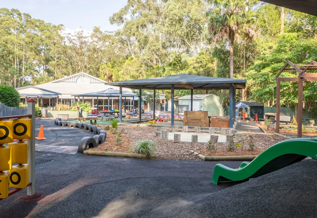 A playground area at day care in the Central Coast with various play structures, including a slide, a timber log yarning circle, and a sandpit.