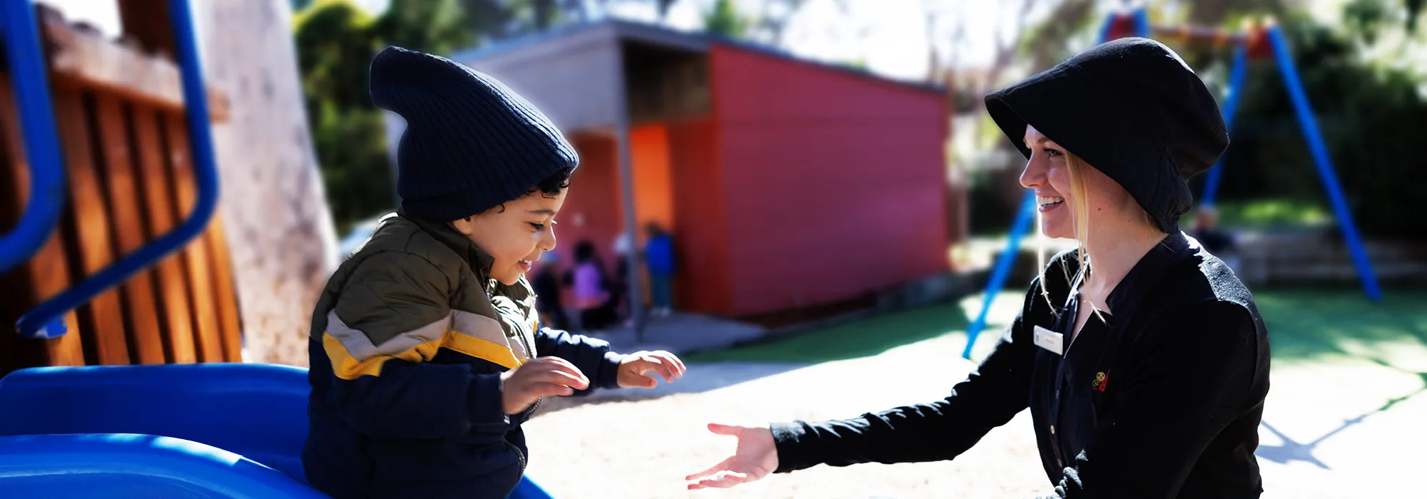 A young child in a beanie slides down a playground slide, reaching out to an adult in a beanie who is waiting to catch them.