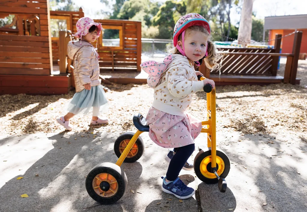 Two young children play outside. One child rides a tricycle while wearing a helmet, and the other child walks nearby. Both are dressed in casual clothing and hats. A wooden structure is visible in the background.