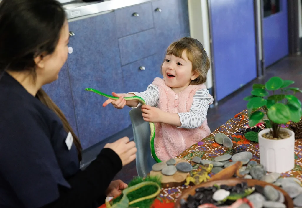 A child in a pink vest shows a green object to an adult seated at a table filled with various small items and a potted plant.