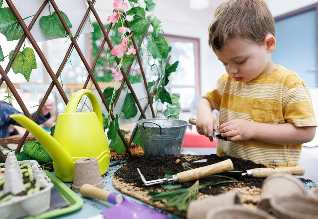 A young boy in a yellow striped shirt is planting seeds in a small indoor garden with various gardening tools and plants around him.