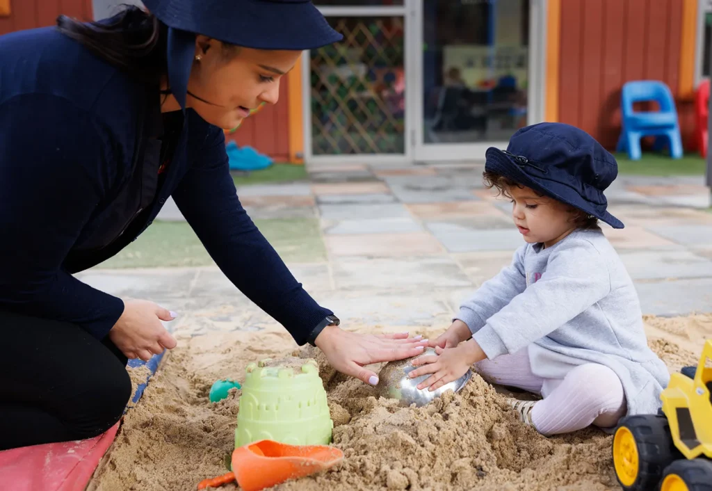 An adult and a child wearing hats are playing together in a sandbox. The child is building in the sand while the adult assists. Plastic toys and a sandcastle mold are also visible.