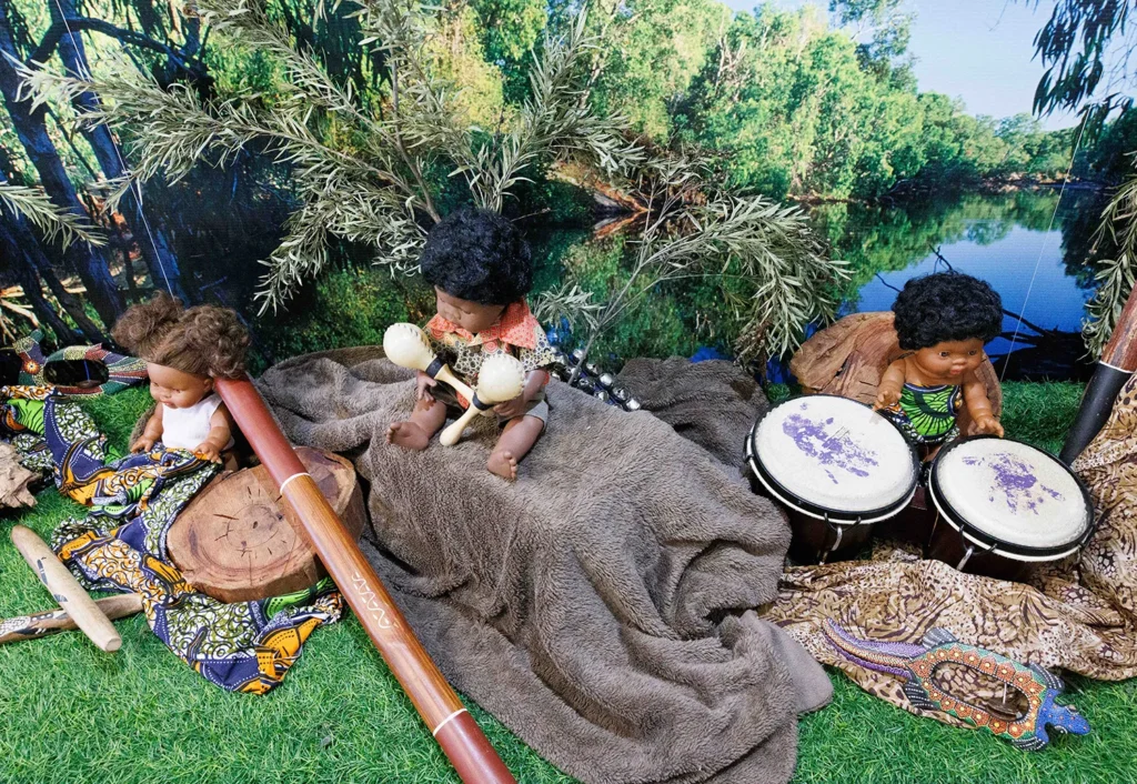 Three dolls sit on a grassy surface surrounded by musical instruments, including a didgeridoo, maracas, and bongos, with a nature backdrop of trees and a pond.