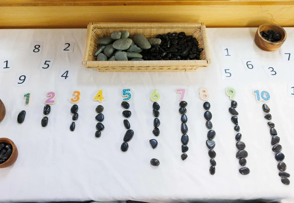 A table with cards displaying the numbers 1 to 10 is accompanied by corresponding groups of small stones beneath each card. A wicker basket and two bowls of stones are also present.