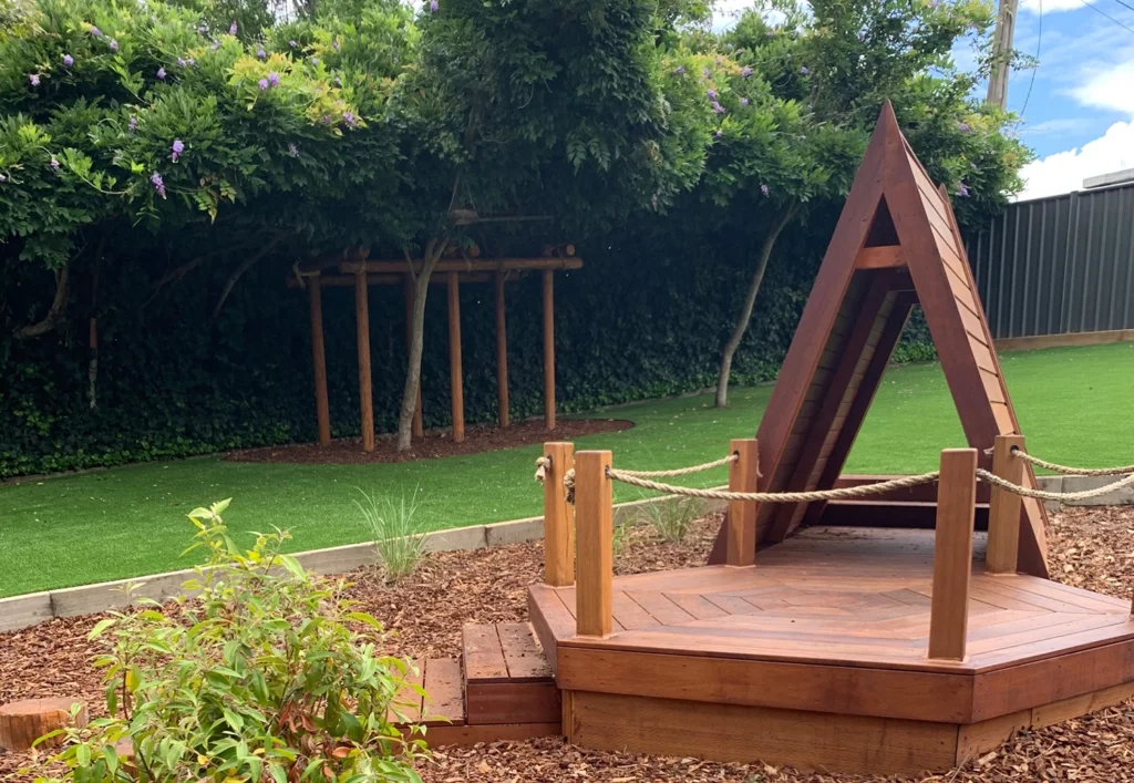 A wooden geometric structure stands on a small platform in a landscaped garden with mulch, artificial grass, and lush greenery in the background.