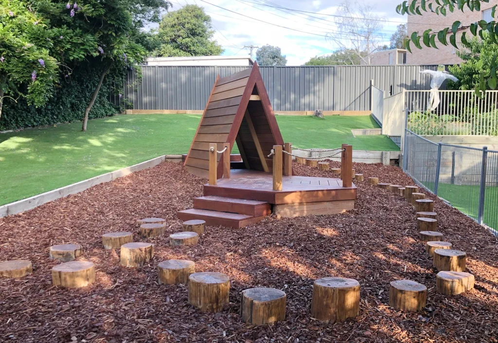 Outdoor playground with a wooden teepee structure and a rope bridge set on mulch. Tree stumps are arranged in a path leading up to the structure. Green trees and a fence are in the background.