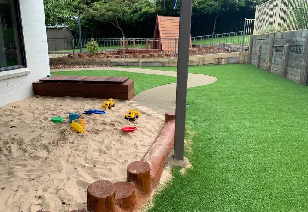 Outdoor playground with sandbox containing toy trucks and shovels, artificial turf, a small playhouse, and a raised wooden garden bed in the background.