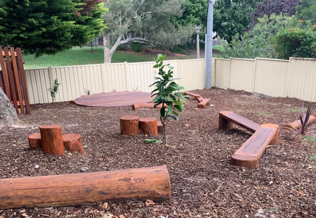 Outdoor seating area with wooden log benches and tree stumps, surrounded by mulch and enclosed by a beige fence. Trees and greenery are in the background.