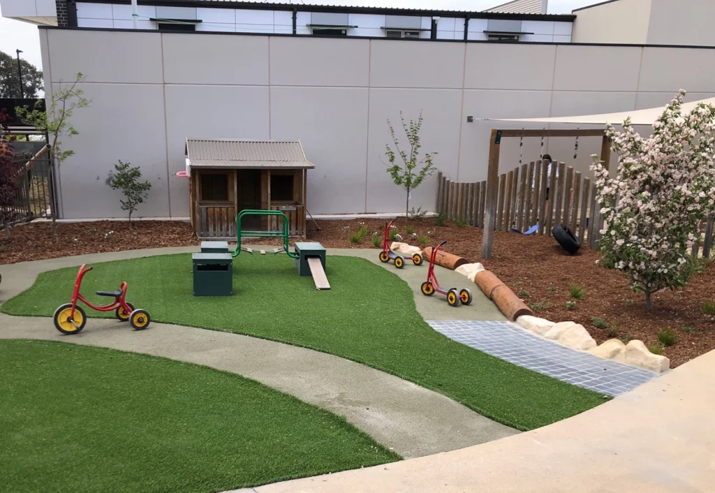 A small playground with artificial grass, a wooden playhouse, several tricycles, a climbing structure, and some flowering plants and trees provides the perfect setting for early learning at this cozy preschool.