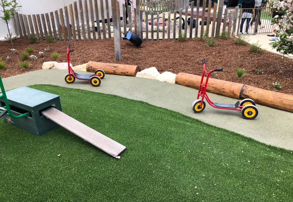 Two children's scooters with yellow wheels and red frames are placed on a path in a playground. The area, ideal for preschool fun, features artificial grass and wooden logs, with a wooden ramp leading to a green structure.