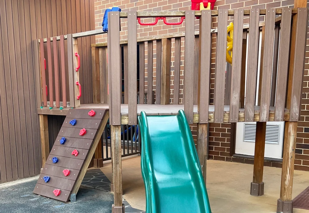 A playground structure perfect for preschool and kindergarten ages features a climbing ramp on the left, a green slide in the center-right, and red and blue handles. This early learning gem is made of wood and set against a brick wall.