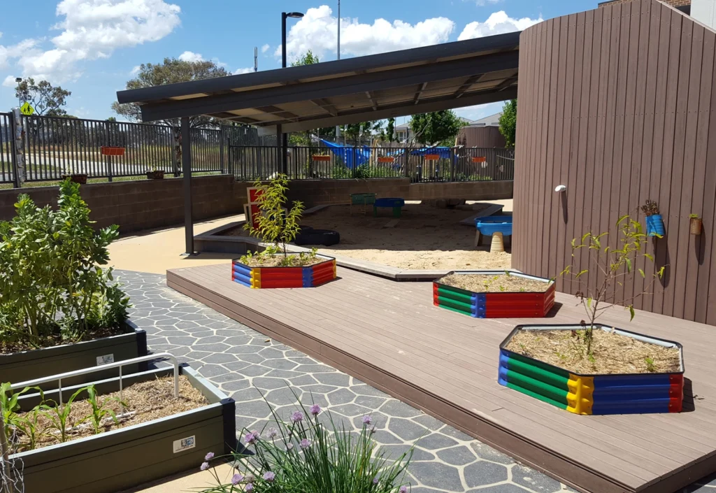 A playground for the kindergarten with a covered sandbox, colorful planter boxes, raised garden beds, and paved walkways. The area is enclosed by a fence, and the playground equipment is visible in the background.