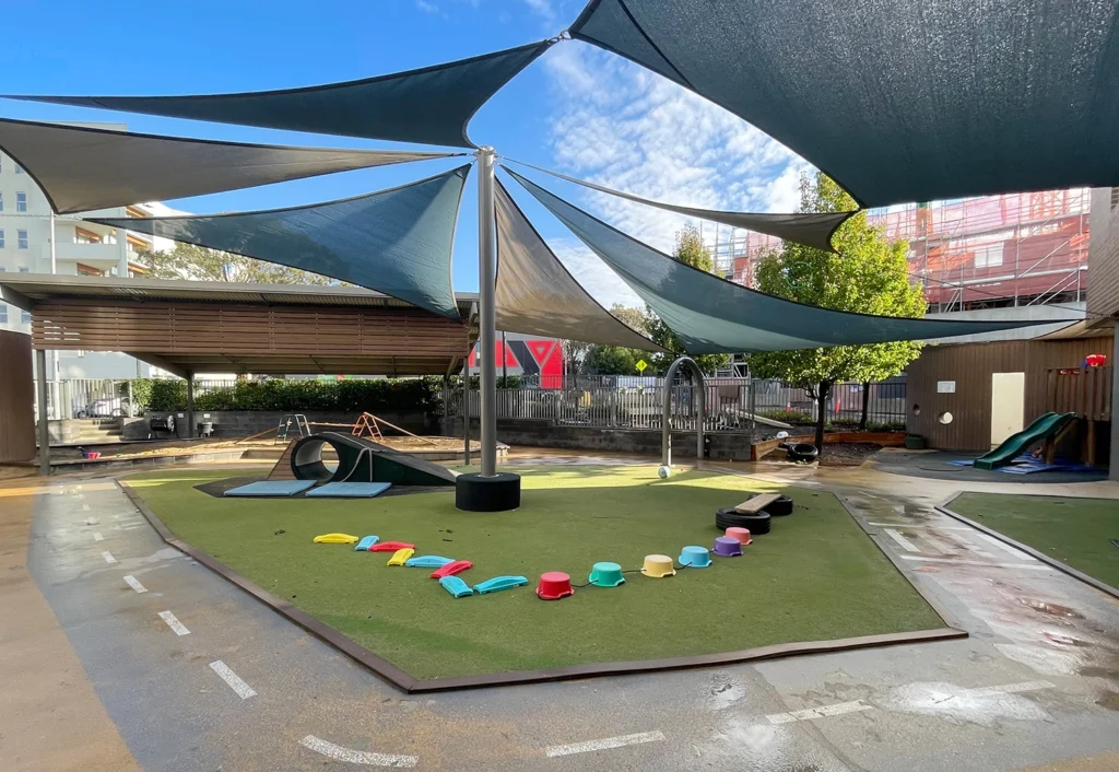 A preschool playground with colorful stepping stones, various play equipment, and large shade sails overhead. The ground is damp, indicating recent rain. Trees and a fence are visible in the background.