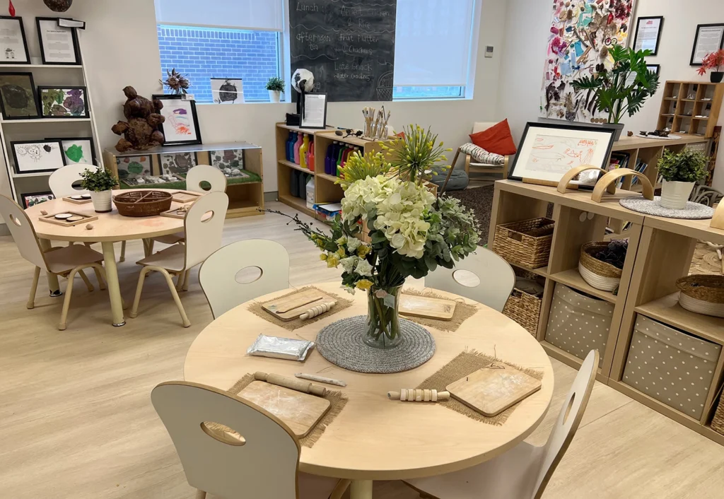 A preschool classroom with round tables set for activities, featuring art supplies and flowers in the center, and various educational materials on shelves against the walls.