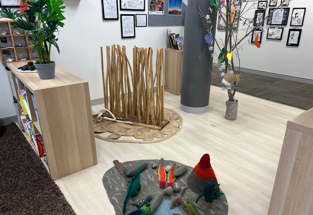 Classroom corner display featuring various art projects, a decorated pole, wooden dividers, low bookshelves with plants, and a rug with a felt scene of aquatic creatures and volcanoes—a perfect setup for any early learning environment or day care center.