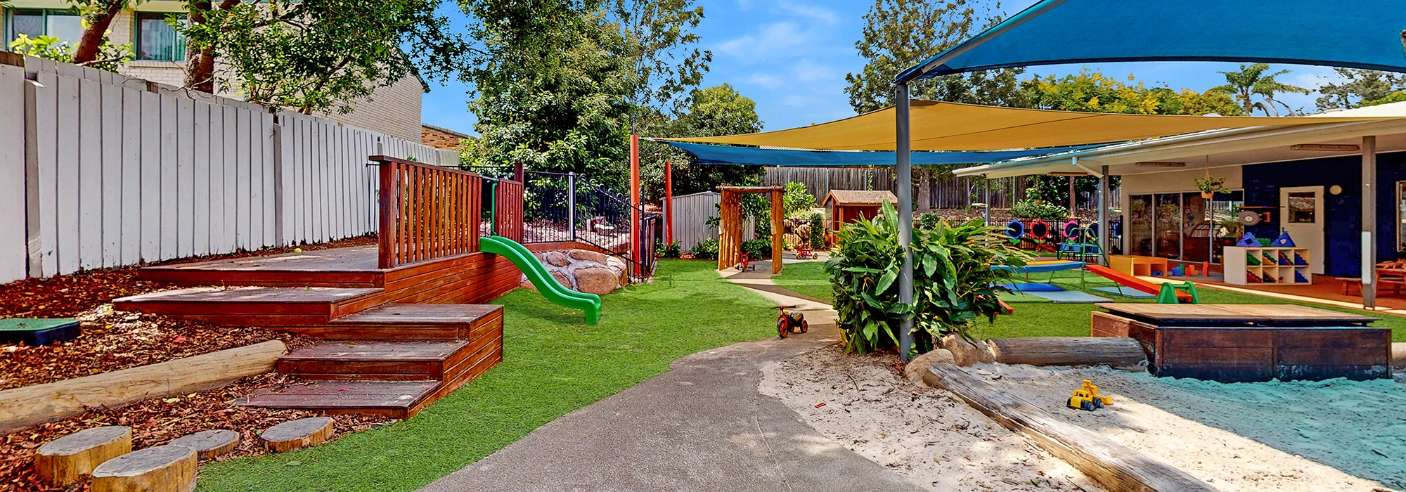 A colorful playground at Busy Bees Underwood childcare features shaded areas, a slide, a sandpit, and various play equipment, all surrounded by trees and a white fence.