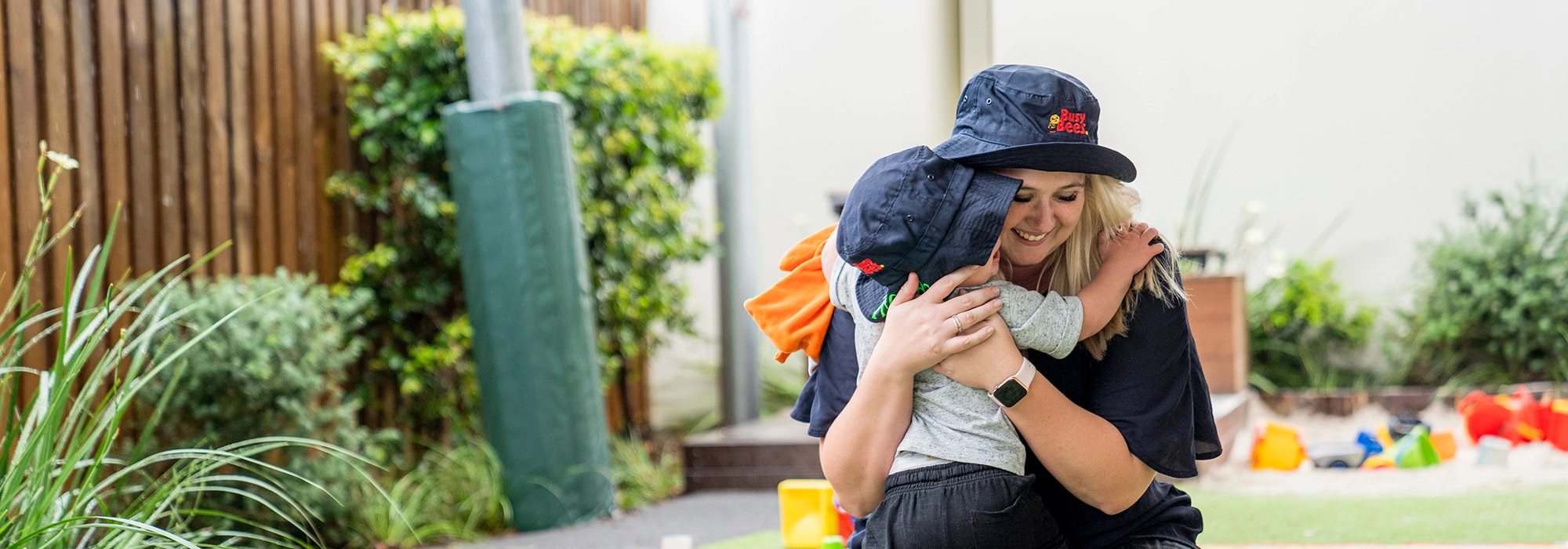 A early learning educator wearing a navy blue hat kneels and hugs a child wearing a matching navy hat in the outside playground of Busy Bees Bellmere daycare.