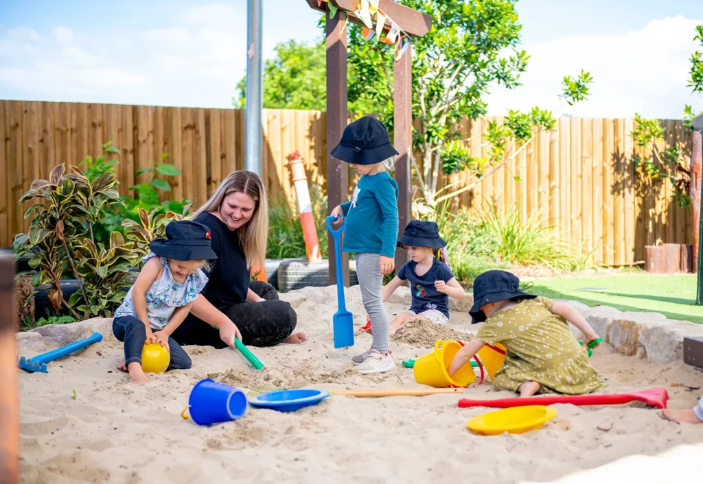 A woman and four young children wearing hats are playing with buckets and shovels in a sandpit in the outdoor playground at Bellmere day care.