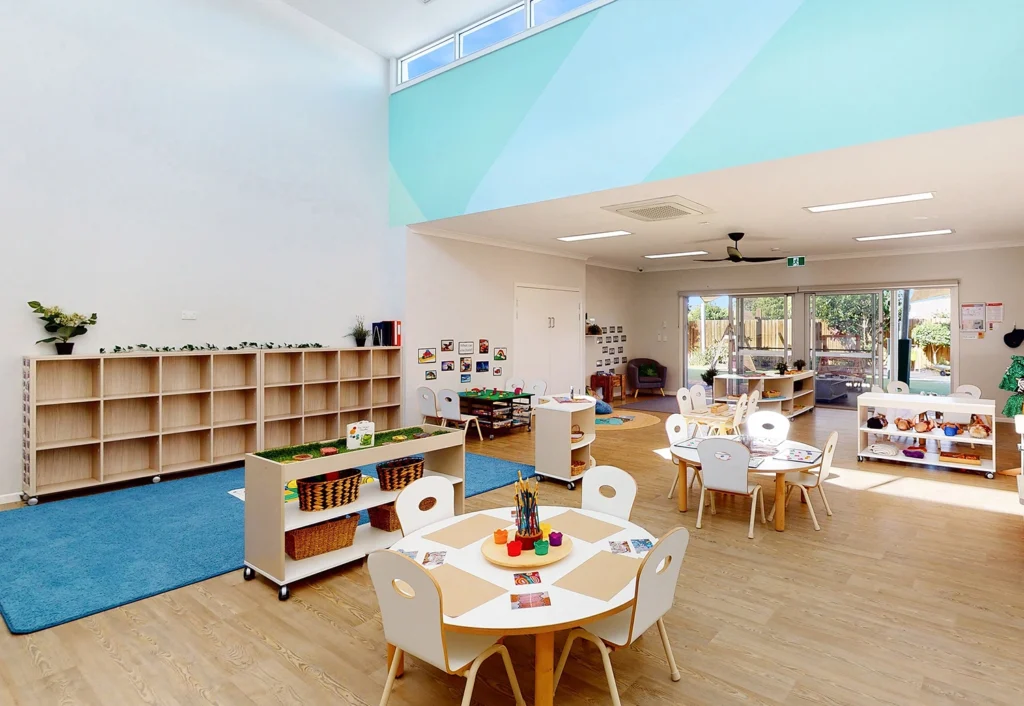 A brightly lit, organized kindergarten classroom at Busy Bees Bellmere with modern furniture, tables, and chairs. Shelves filled with educational materials line one wall, and a blue rug is on the floor. A door leads to an outdoor space.