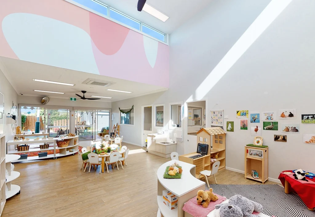 Bright and spacious classroom at Bellmere Childcare with various activity areas, including tables, shelves with toys, a playhouse, and educational wall displays. Large windows and high ceilings provide ample natural light.