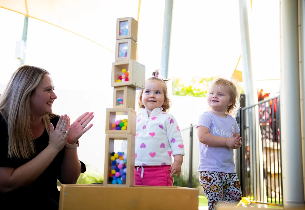 An early learning educator claps her hands while two young children stand next to a tall stack of wooden blocks filled with colorful fabric balls. The scene takes place outdoors at Bellmere day care centre under a shade structure.