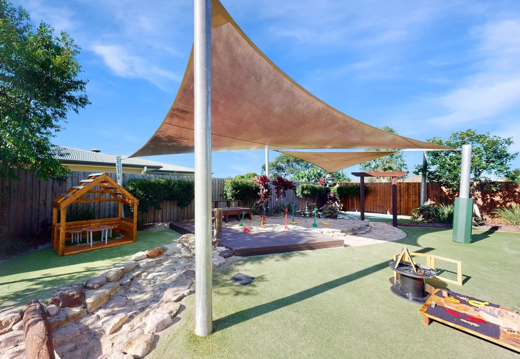 Outdoor playground at childcare for children of Bellmere and Caboolture area. The play area features a sunshade canopy, playhouse, sandpit and various play equipment on a soft, grassy surface surrounded by trees and a fence.