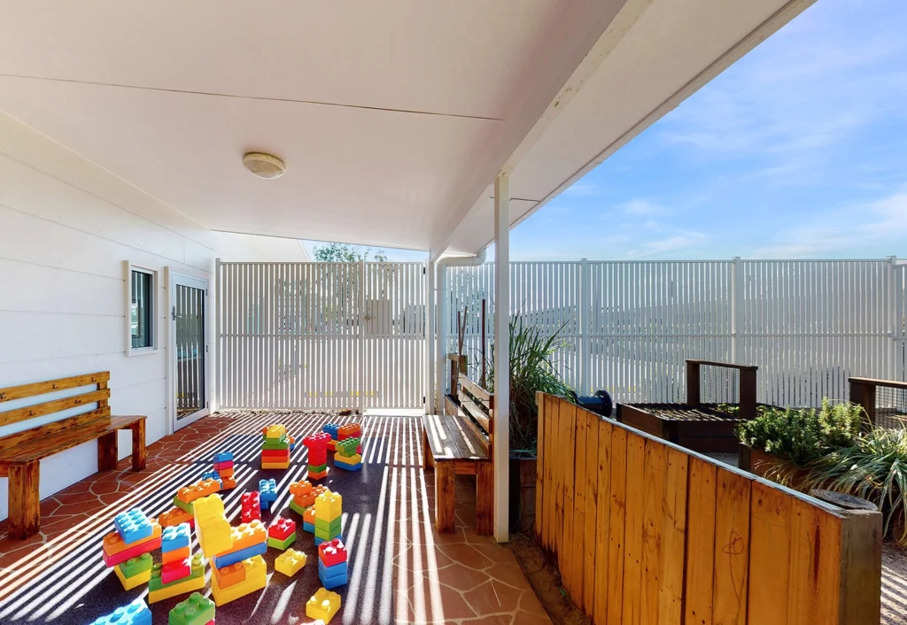 A patio area at Bellmere childcare centre with large colorful building blocks on the floor, wooden benches against the walls, and a fenced perimeter, under a clear blue sky.