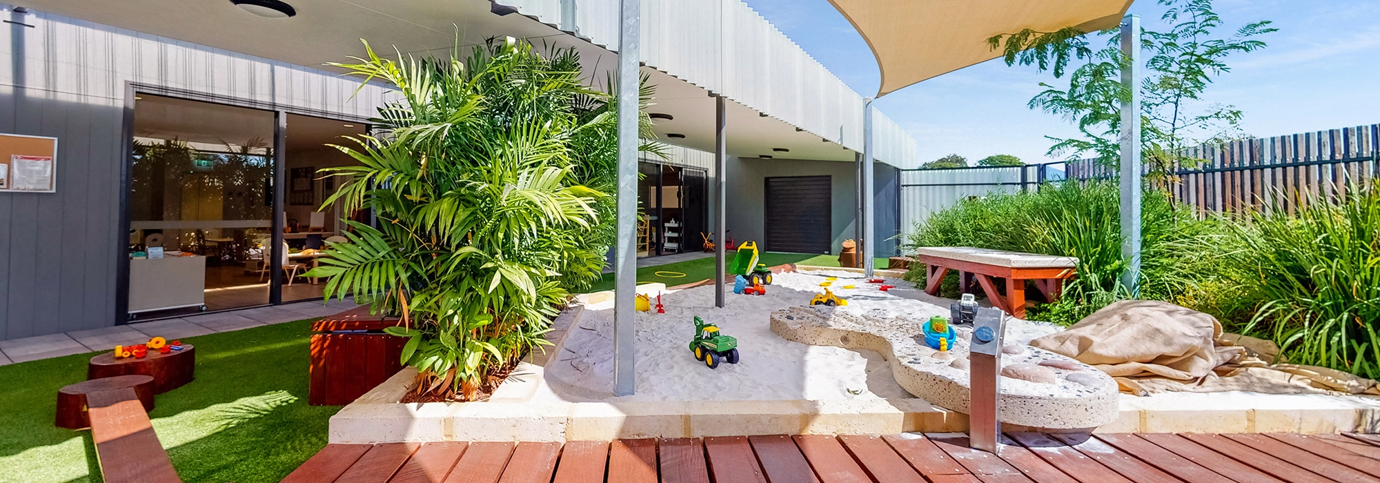 Outdoor play area with a sandpit, covered by a canopy, surrounded by plants. Various toys and a water feature are present. Building with open windows and doors is in the background.