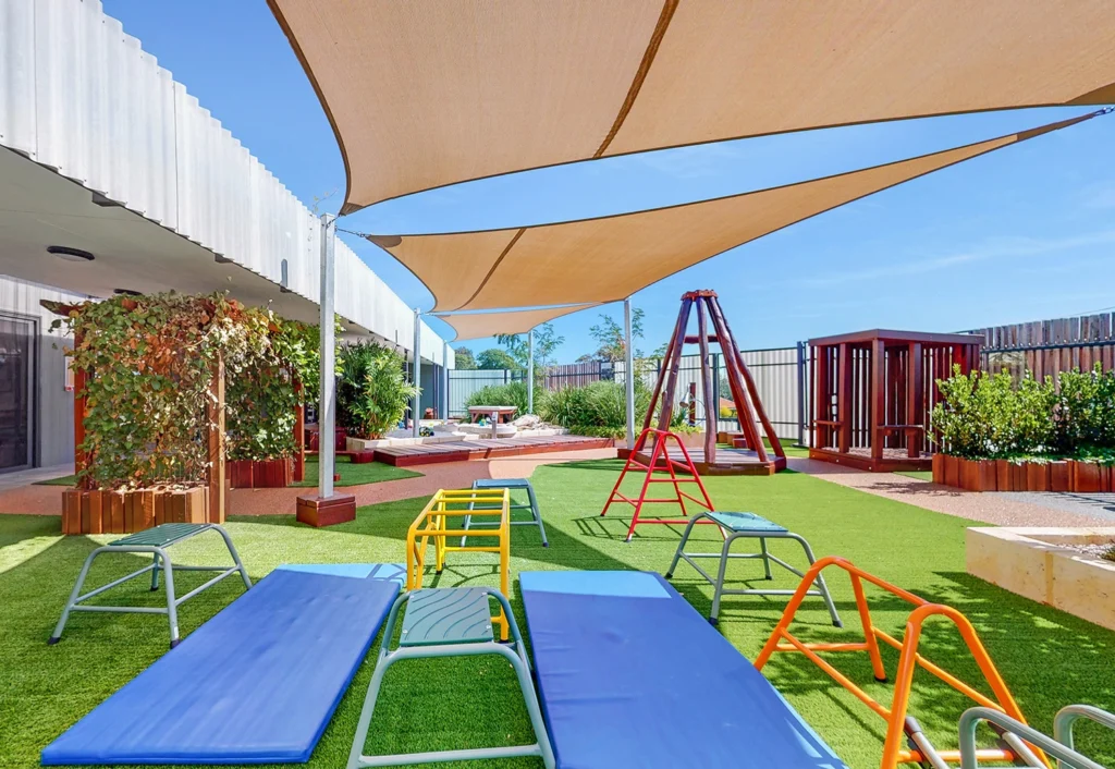 A sunny playground featuring blue and yellow outdoor gym equipment, green artificial grass, shaded areas, a climbing structure, and a garden with a wooden fence in the background.