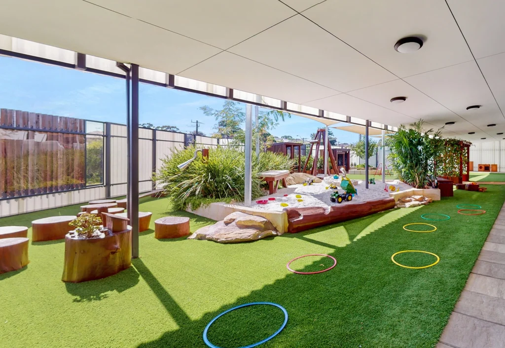 An outdoor play area with artificial grass, wooden stepping stools, a sand pit containing toys, and colorfully arranged hoops on the ground. The space is shaded by a roof and includes greenery.