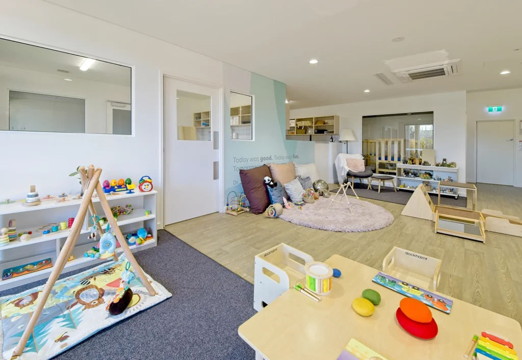 Bright and organized preschool classroom with various toys, activity tables, a play mat, and a cozy reading corner. Shelves are filled with educational materials, and large windows let in ample natural light.