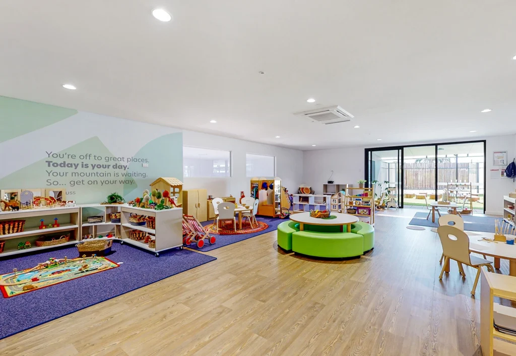 A bright, spacious classroom with various activity stations, an open play area, tables with small chairs, and a motivational quote on the wall. Large windows provide natural light.