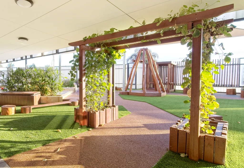 Outdoor play area with artificial turf and a pathway leading under a wooden arbor covered in green vines. In the background, there is a wooden playground structure.