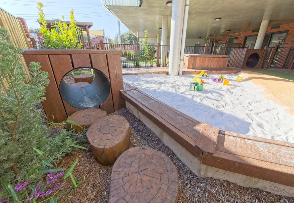 Image of a playground featuring a tunnel, cut tree trunk steps, and a large sandpit with scattered toys. An undercover area and fenced garden are visible in the background.