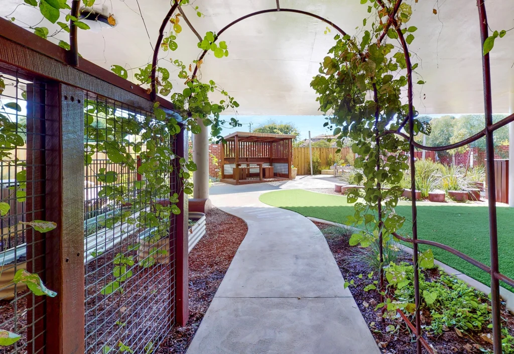 Covered walkway with climbing plants leads to an area with seating under a pergola. The yard features a mix of grass, garden beds, and potted plants.